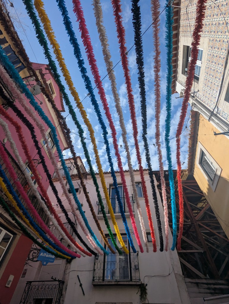 Colorful streamers hanging between buildings in a narrow street, with a blue sky and clouds in the background.