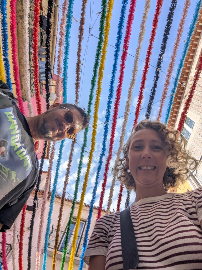 A selfie of two people standing under colorful decorative streamers in a vibrant street, with a blue sky in the background.
