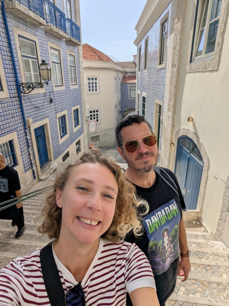 A couple taking a selfie on a cobblestone street lined with blue-tiled buildings and white walls under a clear sky.