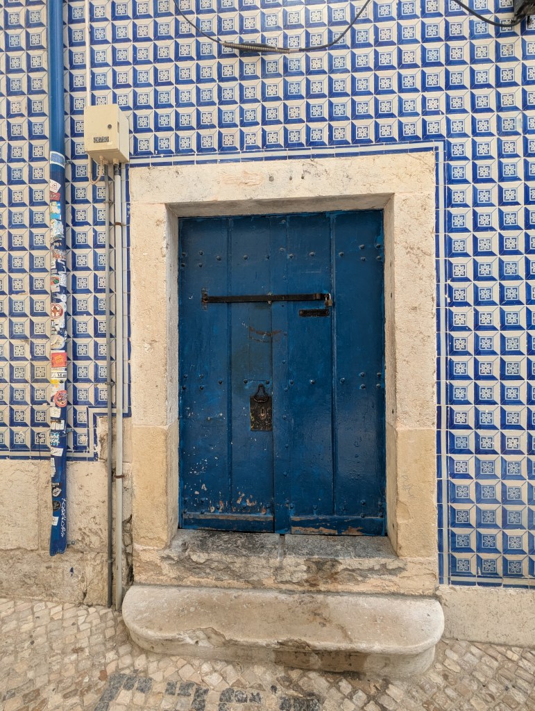 A weathered blue wooden door with a metal latch, set in a stone wall adorned with blue and white patterned tiles.