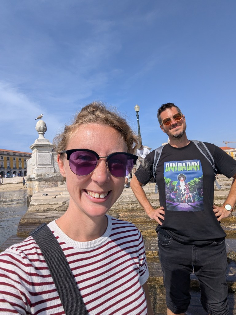 Two people smiling for a selfie outdoors near a body of water, with one person wearing sunglasses and a graphic t-shirt.