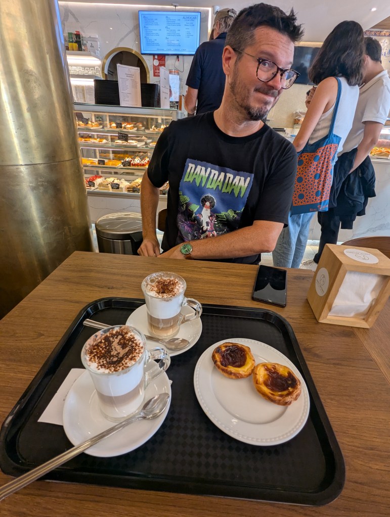 A man with glasses is sitting at a café table, smiling playfully. In front of him are two glasses of layered coffee drinks topped with cream and cocoa, and two Portuguese custard tarts on a white plate. A display case filled with pastries is visible in the background.