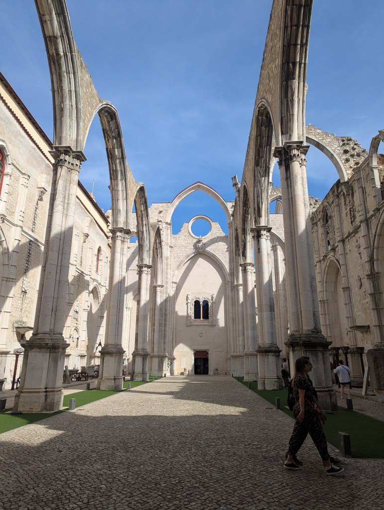Interior view of a historic church ruin with tall stone arches and a clear blue sky above. People are walking along a cobblestone path, with greenery visible on the ground.