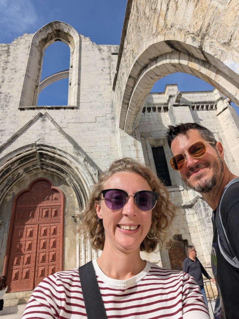 A selfie of two people in front of a historic, partially-ruined church with an arched entrance and large ornate door. Clear blue sky is visible above.