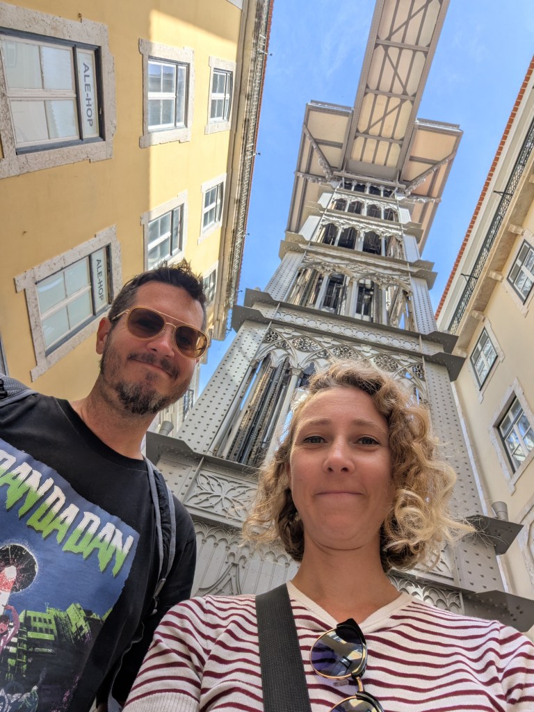 A selfie of a man and a woman standing in front of the Elevador de Santa Justa in Lisbon, Portugal, with surrounding buildings and a clear blue sky.
