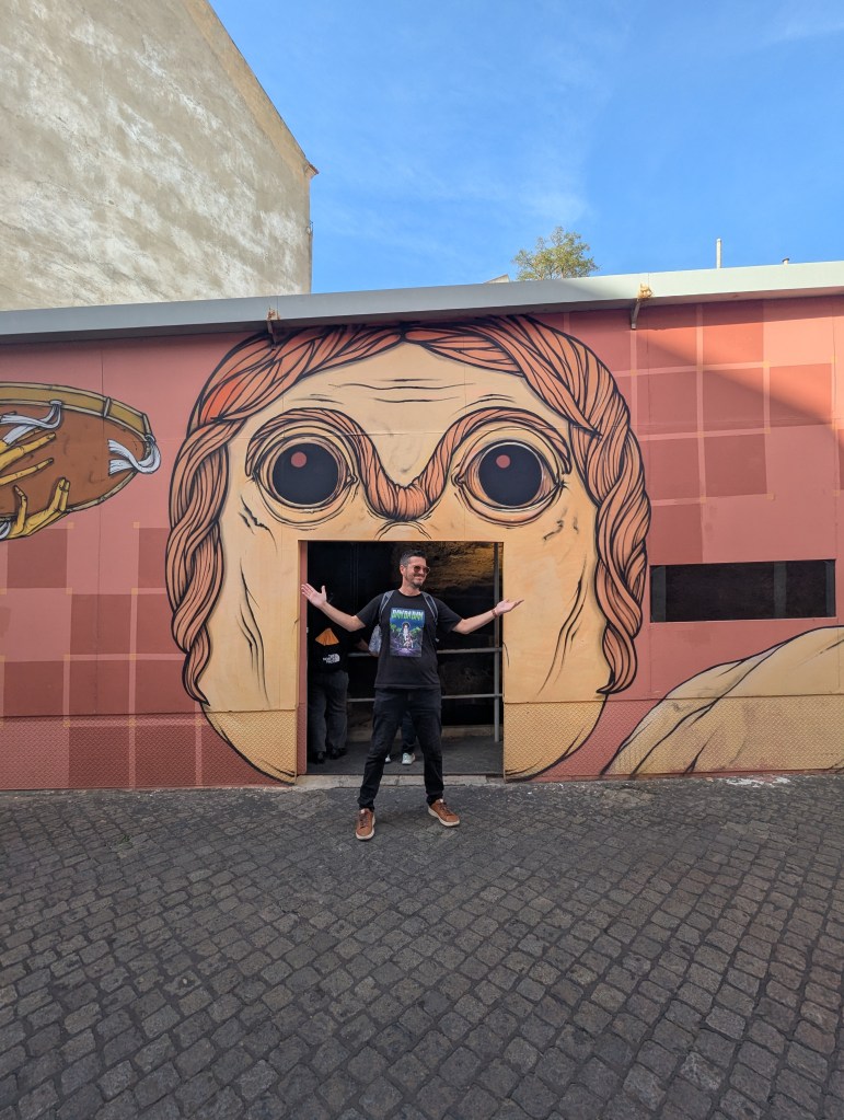 A man stands in front of a colorful mural depicting a large face with exaggerated features, with his arms outstretched in a welcoming gesture. The background features a clear blue sky and a textured surface.