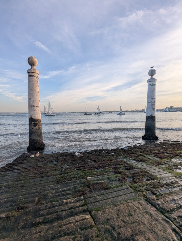View of a waterfront with two tall pillars, sailing boats in the background, and a sandy, moss-covered shore under a cloudy sky.