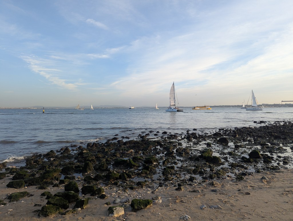 A coastal scene with sailboats on a calm sea, rocky shoreline in the foreground, and a clear sky with wispy clouds.