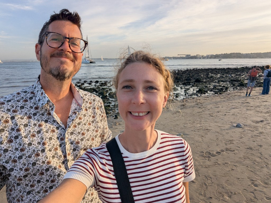 A man in glasses and a patterned shirt and a woman with curly hair in a striped shirt smile for a selfie on a beach, with a rocky shoreline and a sailboat in the background under a clear sky.