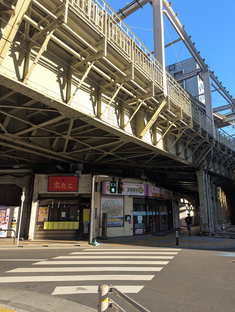 A street scene under a large elevated bridge featuring a restaurant with red signage and various shops. A pedestrian crossing is visible in the foreground, and traffic lights are seen nearby.
