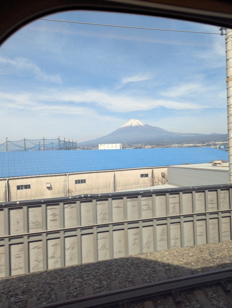 View of snow-capped Mount Fuji from a train window, with industrial buildings and blue roofs in the foreground under a clear sky.