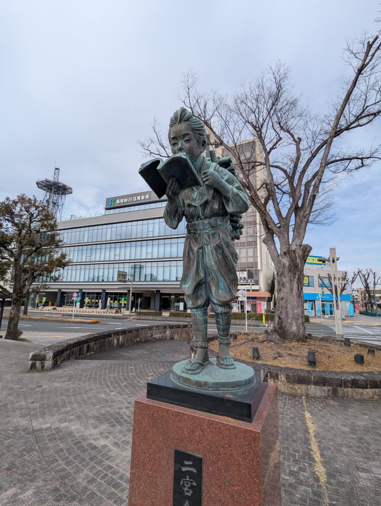 A bronze statue of a young child reading a book, standing on a stone pedestal, with a tree and modern buildings in the background.