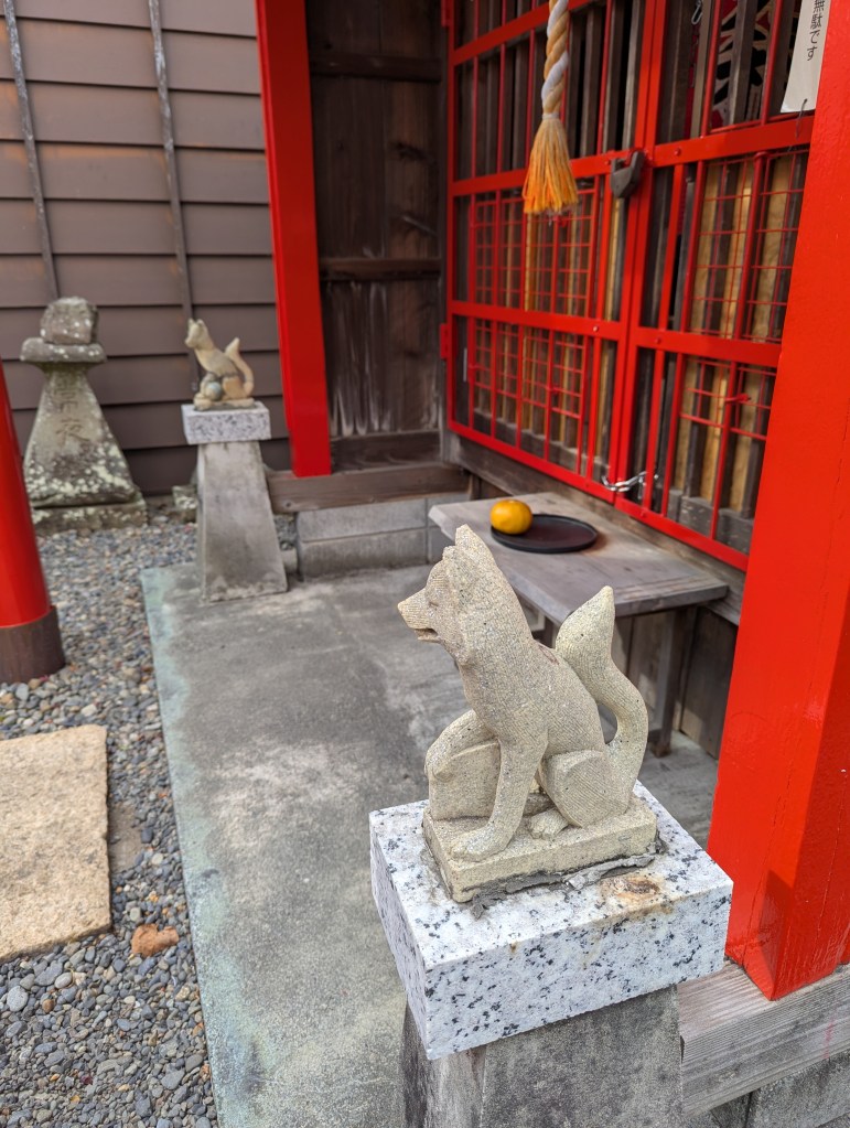 Stone fox statues at the entrance of a traditional Japanese shrine, with a wooden structure and red gates in the background.