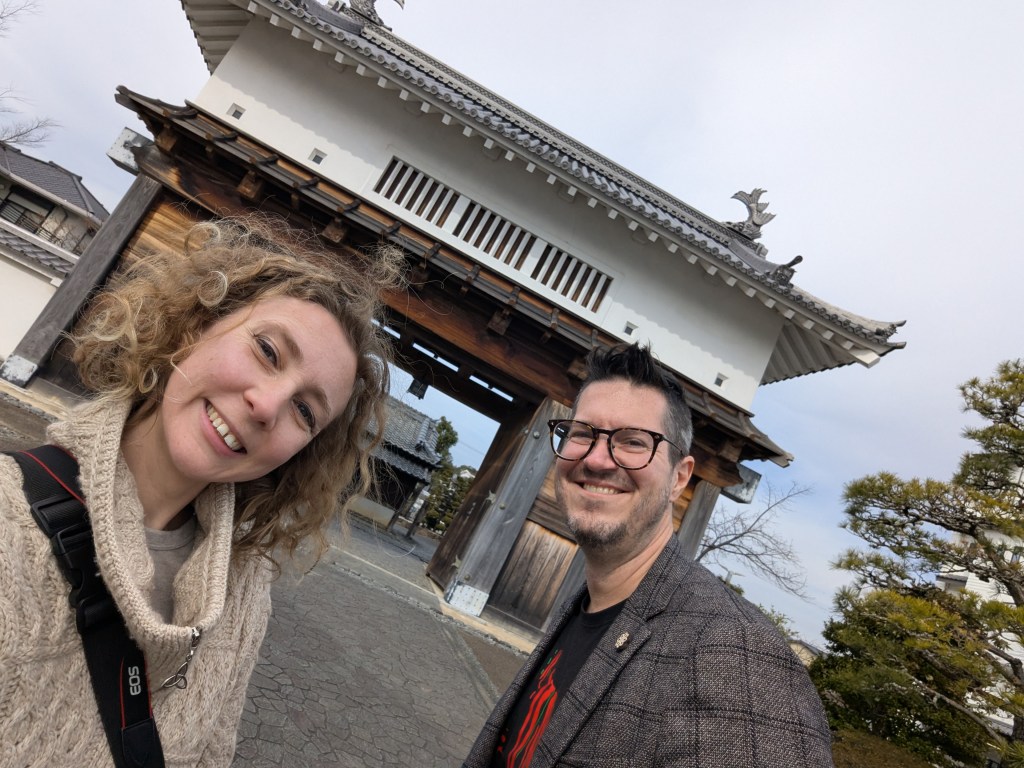 A smiling woman and a man pose for a selfie in front of a traditional Japanese gate.
