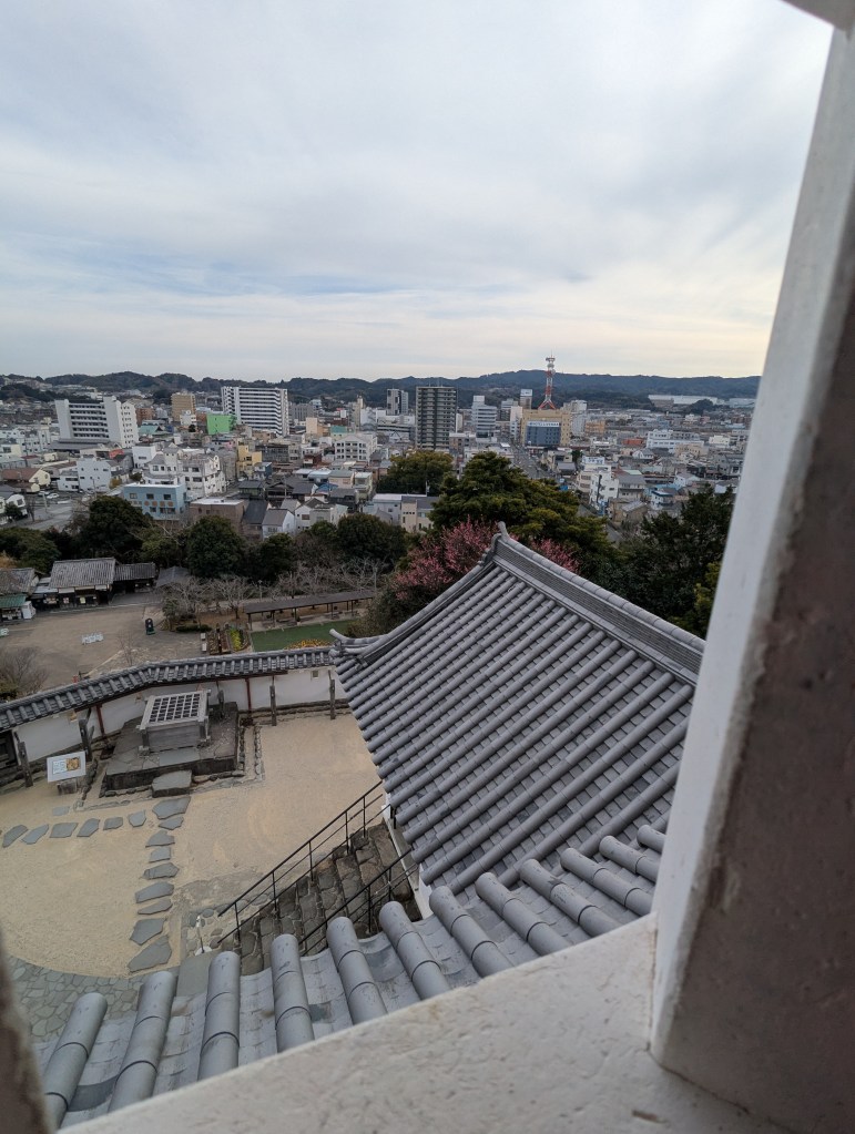View from a window showing traditional tiled roofs and a cityscape with modern buildings in the background under a cloudy sky.
