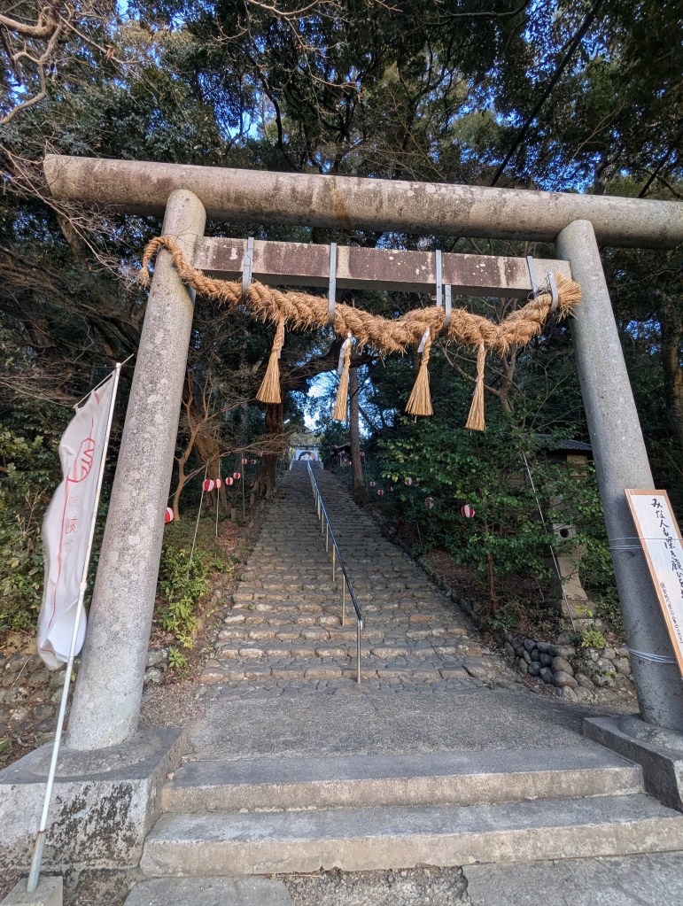 A stone torii gate adorned with a rope hangs over a set of stairs leading up a path, surrounded by lush greenery.