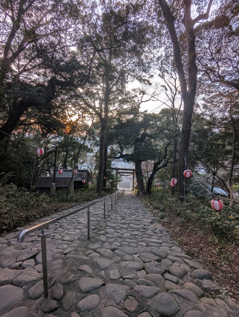 A stone pathway leading through a forested area, flanked by trees and traditional lanterns, leading to a torii gate in the distance under a soft sunset.