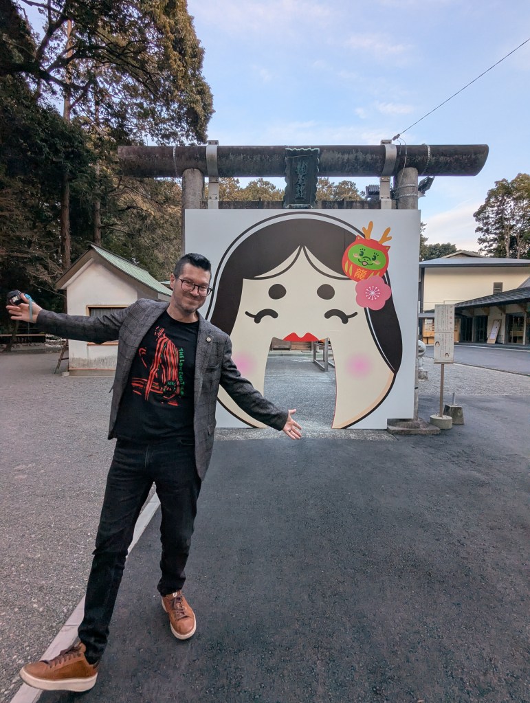 A person stands with arms outstretched in front of a large, cartoonish gate featuring a stylized face with a playful expression, surrounded by trees and buildings.