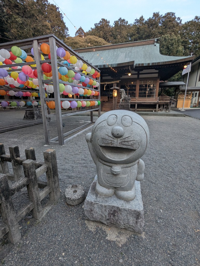 Stone statue of a cartoon character in a shrine courtyard, with colorful lanterns and traditional Japanese architecture in the background.