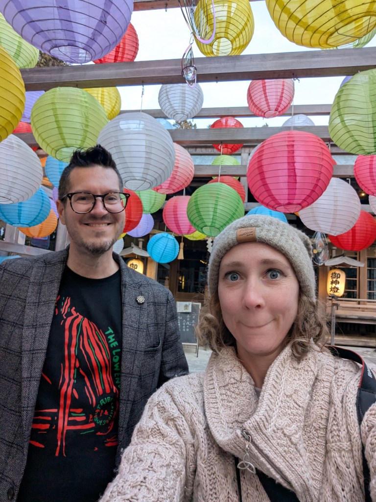 A selfie of two people standing under colorful lanterns in a vibrant outdoor setting. One person is wearing glasses and a blazer, while the other is dressed in a cozy sweater and beanie.