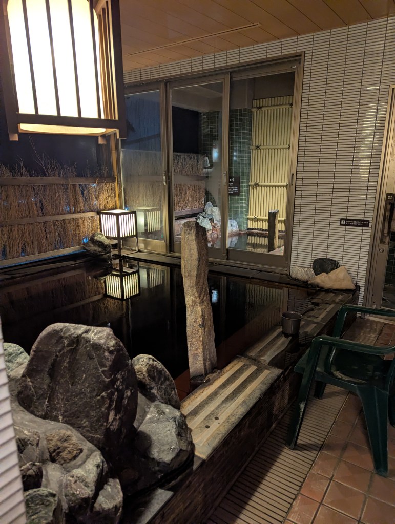 A serene view of an outdoor hot spring area featuring dark water, stone decorations, and a glowing lantern, with a glimpse of a seating area through glass doors.