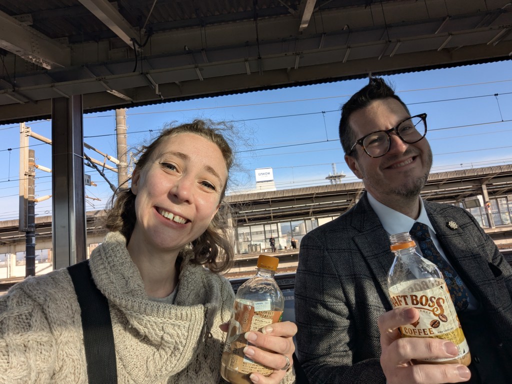 Two people sitting at a train station, smiling and holding bottles of iced coffee. The background shows train tracks and a clear blue sky.