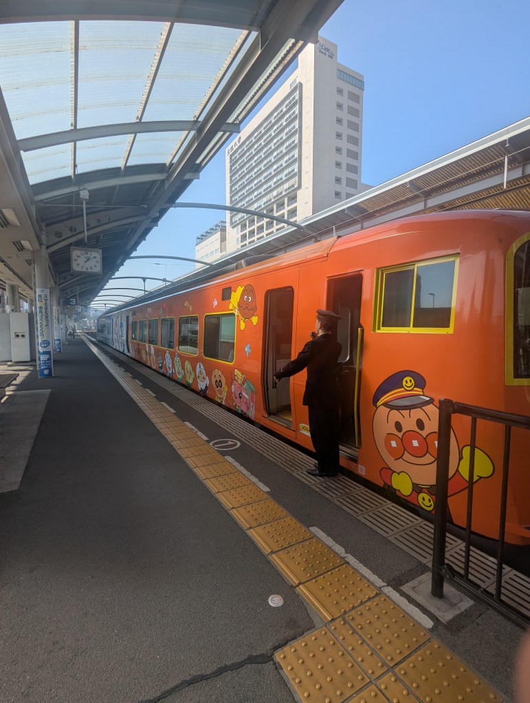 A vibrant orange train decorated with cartoon characters at a station, with a train conductor standing at the door, ready to assist passengers.