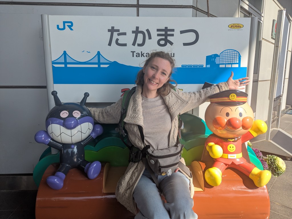 A smiling woman poses with two colorful character sculptures at a train station, featuring a purple cartoon character with a broad grin on her left and a round, orange character wearing a captain's hat on her right. The background shows a sign that reads 'Takamatsu' in Japanese.