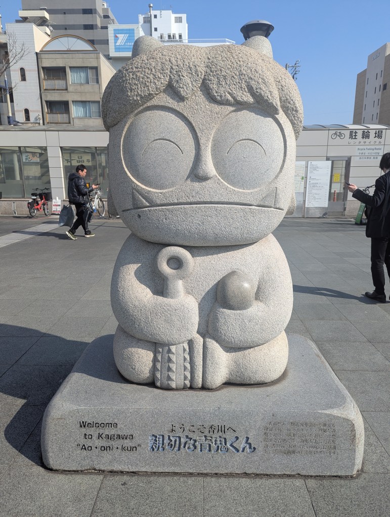A stone statue of a character named 'Aooni-kun' in Kagawa, with a welcoming inscription in both English and Japanese.