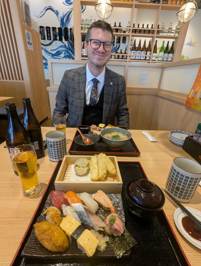 A man in a suit sitting at a restaurant table with a wide smile, surrounded by various Japanese dishes including sushi and tempura, along with beverages like beer and tea.