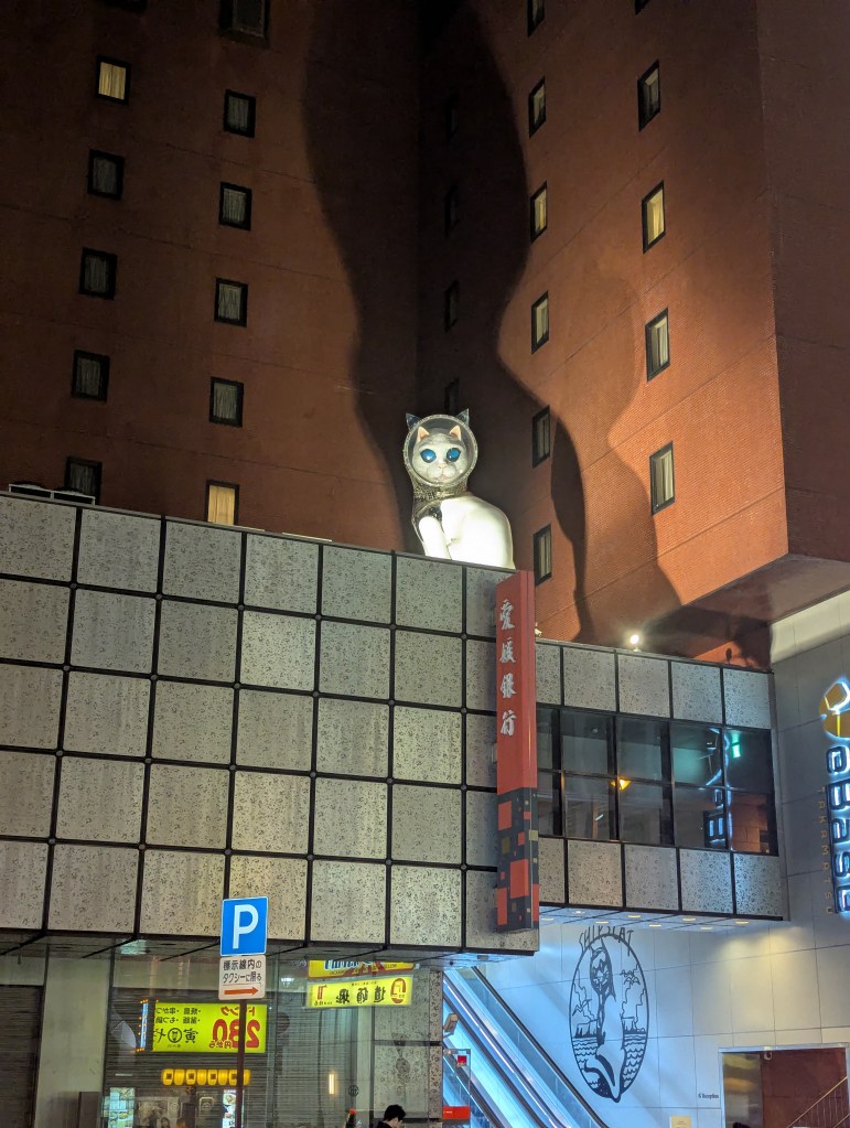 A large, cartoonish white cat with blue eyes sits atop a building, casting a shadow on the wall of a nearby structure. The scene is set at night with illuminated windows and signs visible.